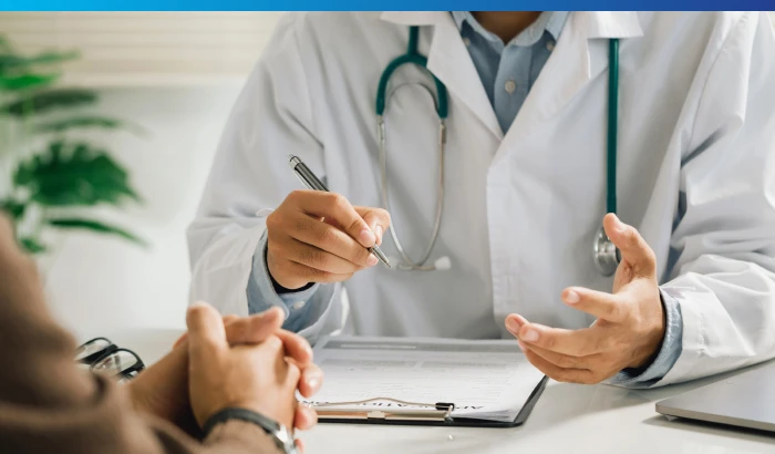 Male physician in white lab coat sitting at desk holding pen and writing on a clipboard while talking to a patient.